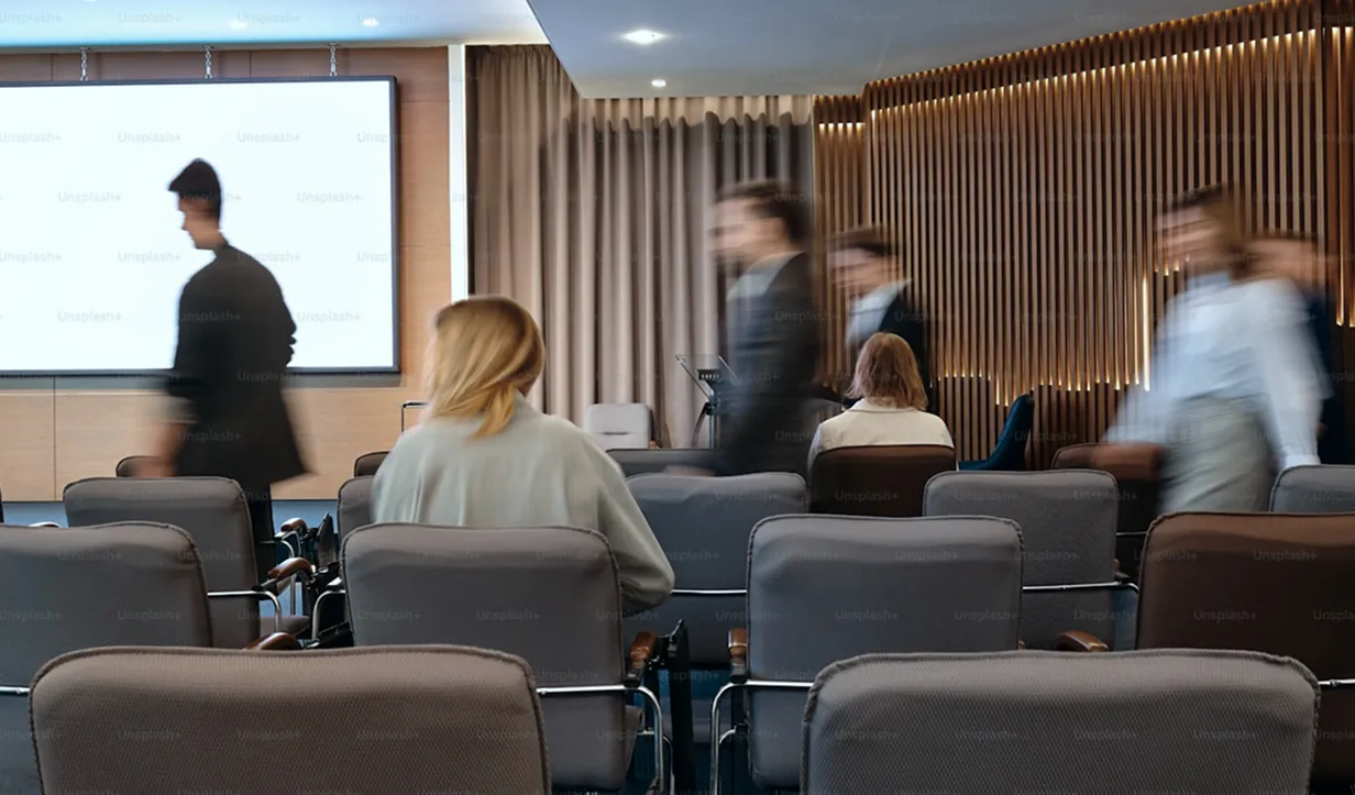 People walking past seated attendees in a conference room with gray chairs and a large screen.