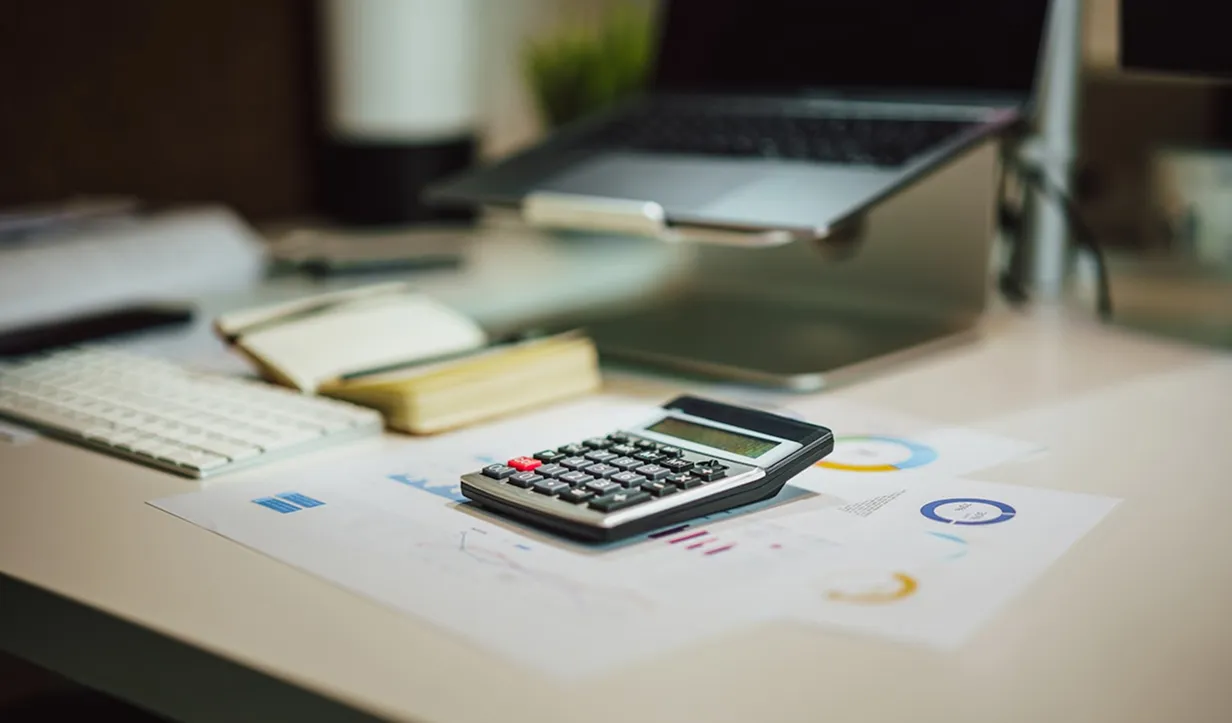 Calculator resting on financial documents on a desk with a keyboard, notebook, and laptop in the background.
