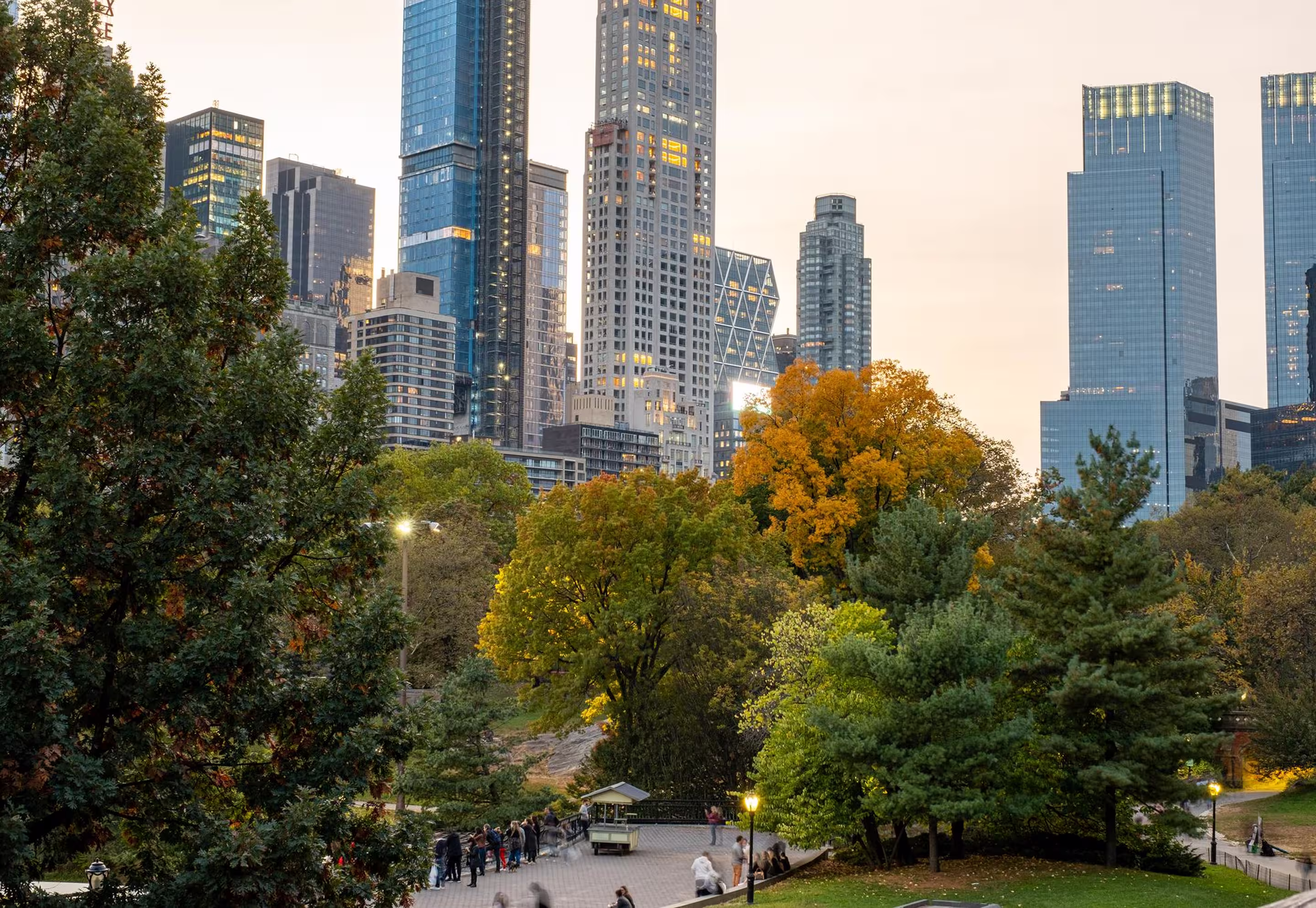 City park in autumn with green and orange trees and a paved walking area with people, skyscrapers in the background.