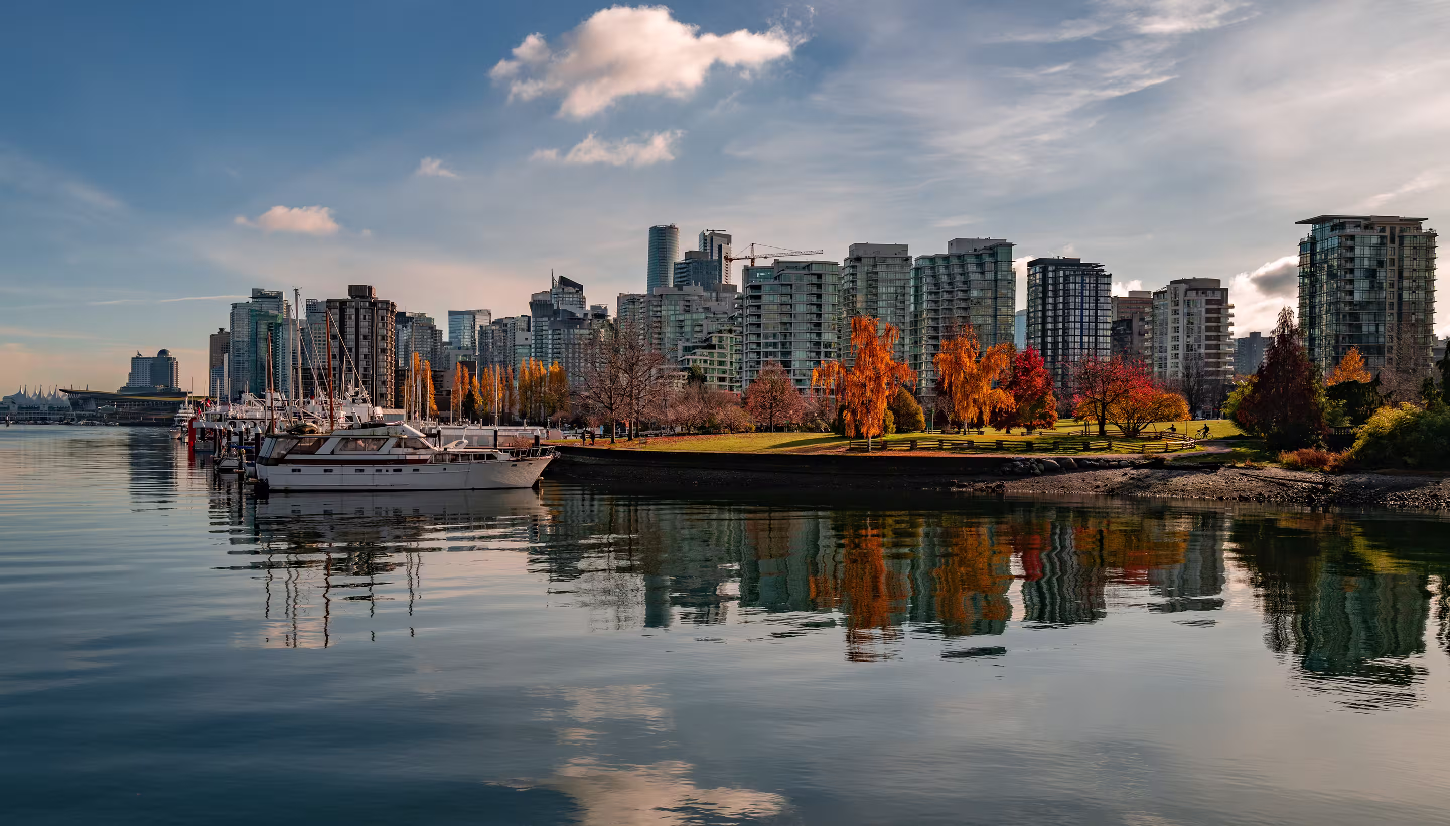 Calm waterfront with docked boats, autumn trees, and modern city skyline under a partly cloudy sky.