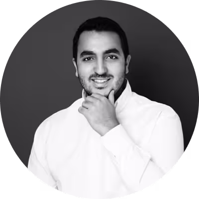 Smiling man with short hair and beard wearing a white shirt with one hand resting on his chin, posed against a dark background.