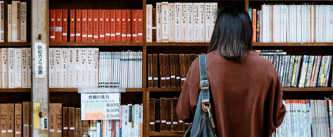 Adult learner browsing tall shelves of books in a bookstore, continuing self-directed study