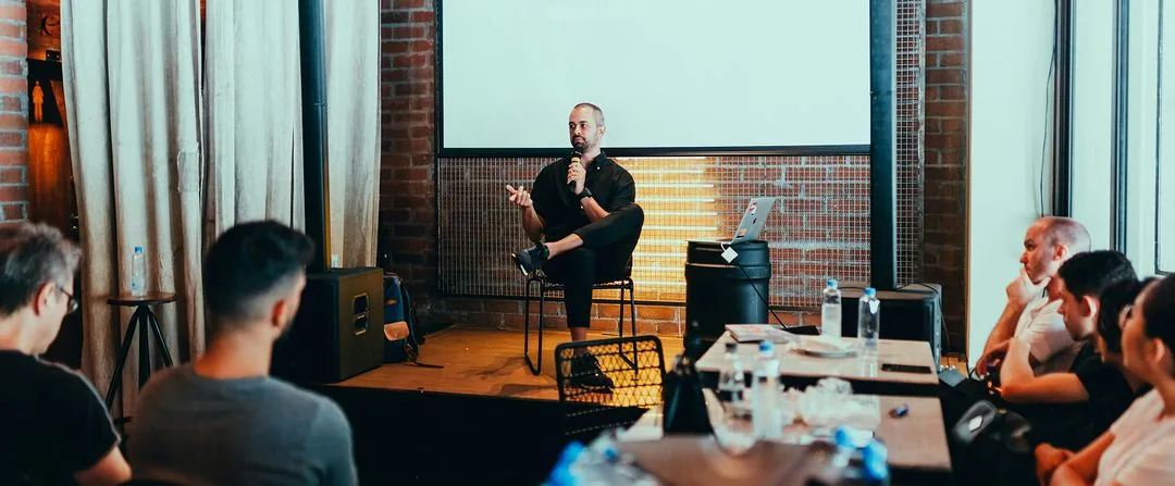 Speaker seated on a stool with a microphone addressing a small audience in a conversational talk