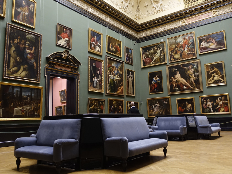 Interior of an art museum room with classical paintings hanging on green walls and several gray cushioned benches on wooden flooring.