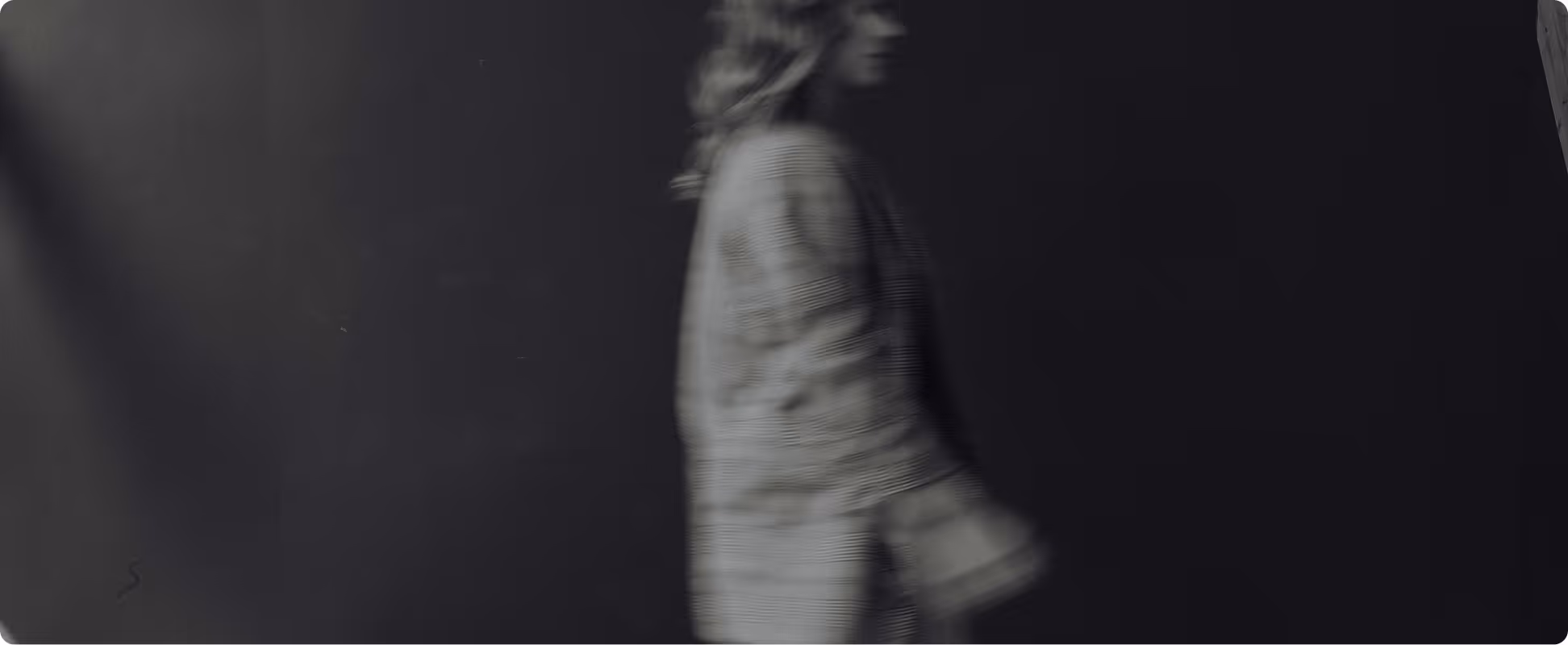 Blurred side profile of a person with long hair wearing a checkered coat against a dark background.