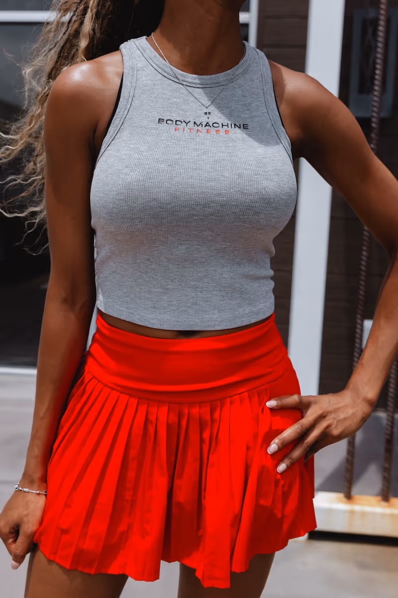 Woman wearing a gray sleeveless crop top with 'BODY MACHINE FITNESS' text and a red pleated skirt, standing with hand on hip.
