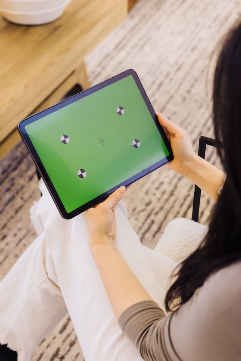 Person holding a tablet with a green screen and calibration markers while sitting on a chair indoors.