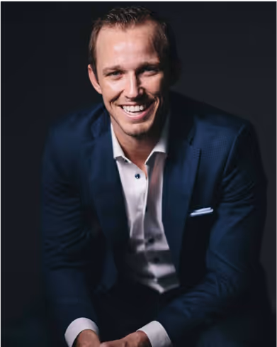 Smiling man wearing a dark blue suit and white shirt against a black background.