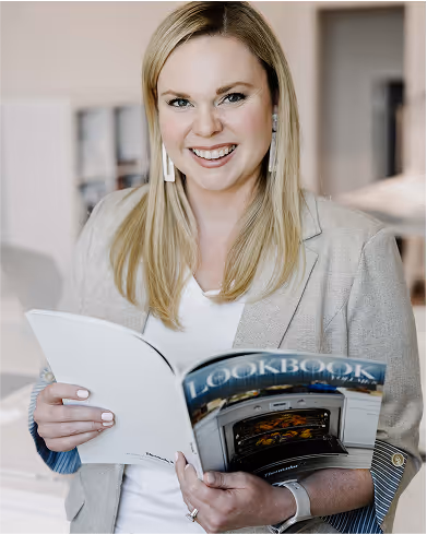 Smiling woman with long blonde hair holding an open cookbook in a bright kitchen setting.