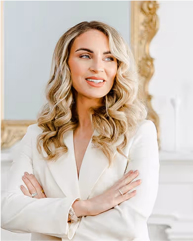 Smiling woman with long blonde wavy hair wearing a white blazer, standing with arms crossed.
