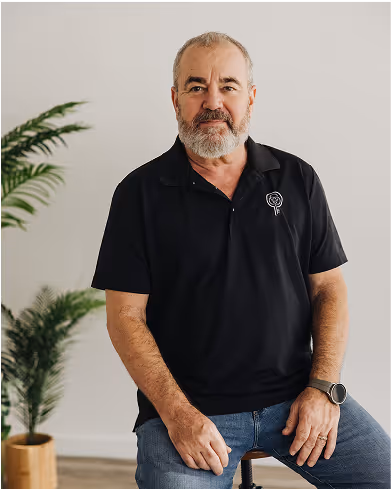 Middle-aged man with gray beard wearing a black polo shirt and jeans, sitting on a stool with a plant in the background.