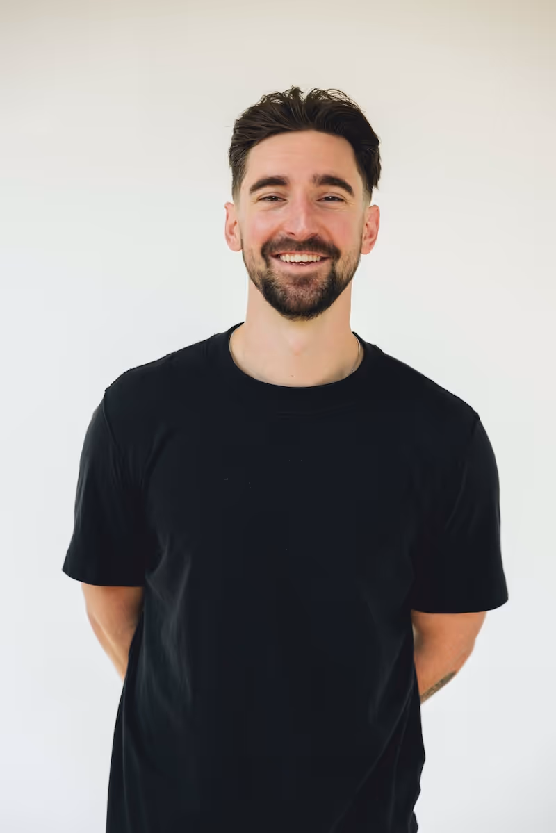 Smiling man with dark hair and beard wearing a black t-shirt standing against a plain light background.