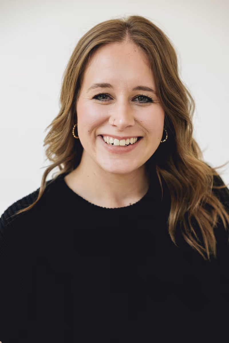 Smiling woman with wavy light brown hair wearing hoop earrings and a black top against a plain light background.