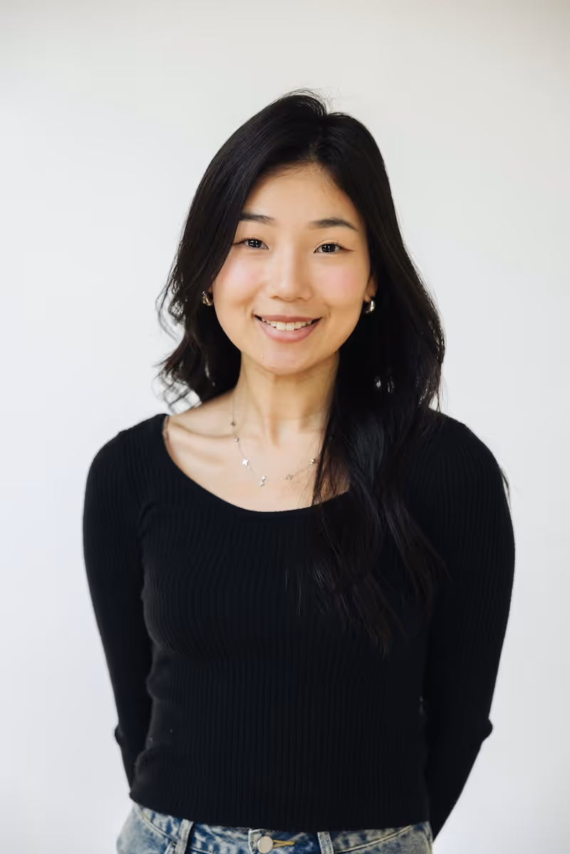 Smiling young woman with long black hair wearing a black sweater and denim jeans against a plain white background.