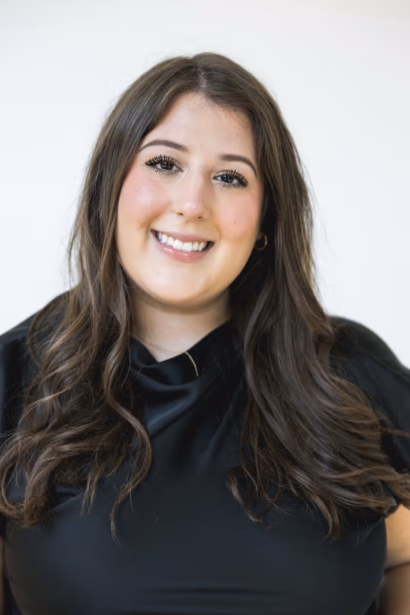 Smiling woman with long wavy brown hair wearing a black top against a plain light background.