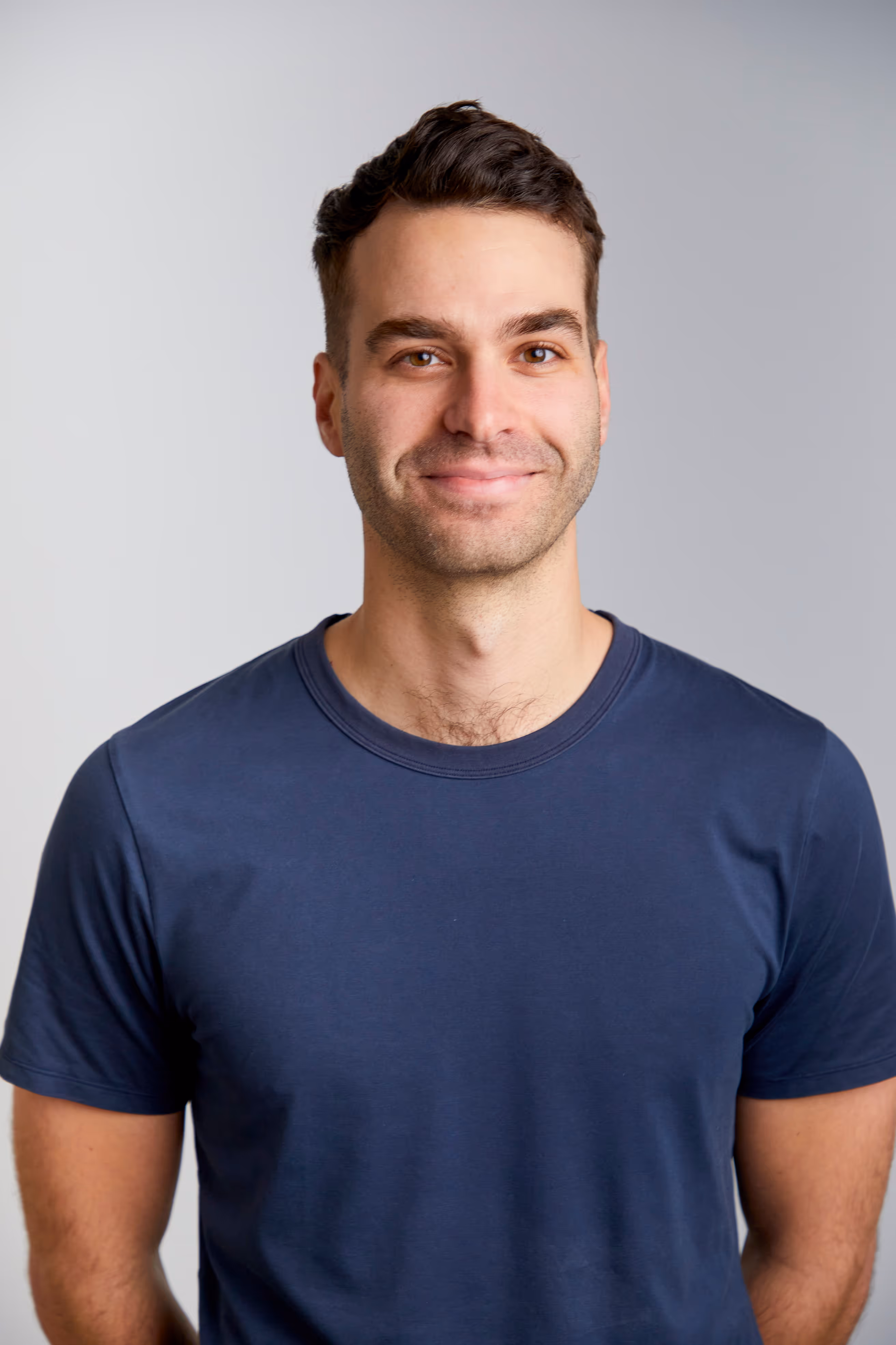 Smiling man with short dark hair wearing a navy blue t-shirt against a plain light gray background.