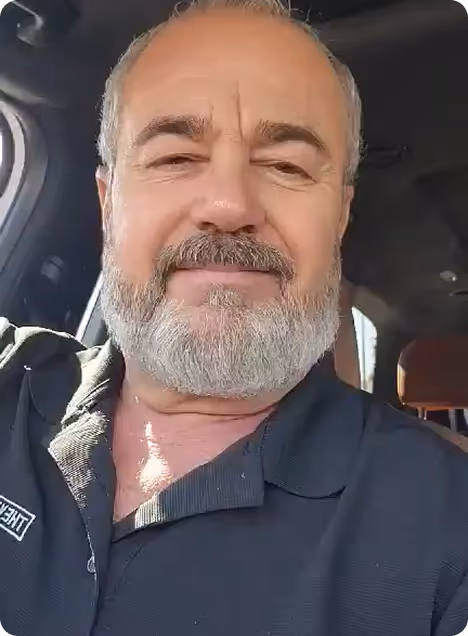 Middle-aged man with a gray beard and mustache smiling inside a car, wearing a dark collared shirt.