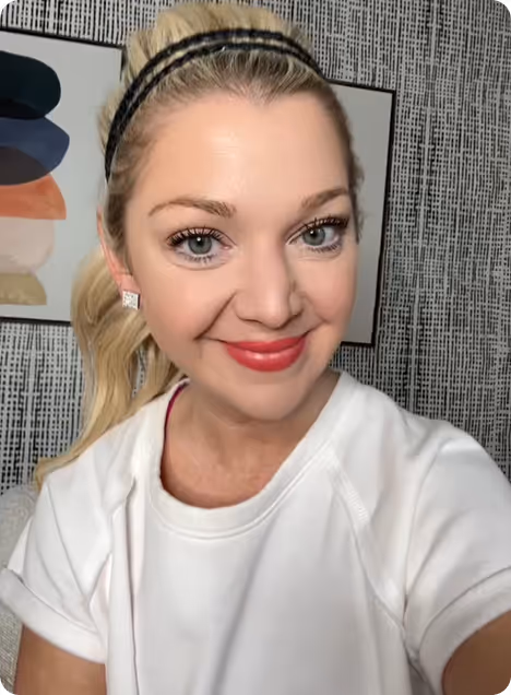 Smiling woman with blonde hair in a ponytail wearing a white shirt and black headband, posed indoors with abstract art in the background.