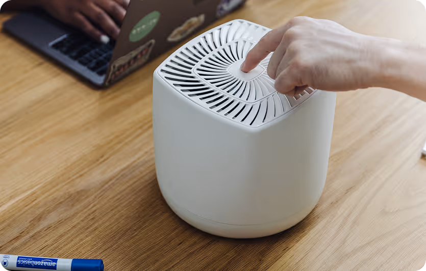 Person pressing the top button of a white small air purifier on a wooden table next to a blue marker and a laptop.