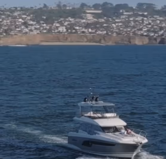 White motor yacht cruising on calm blue water with a coastal city and cliffs in the background.