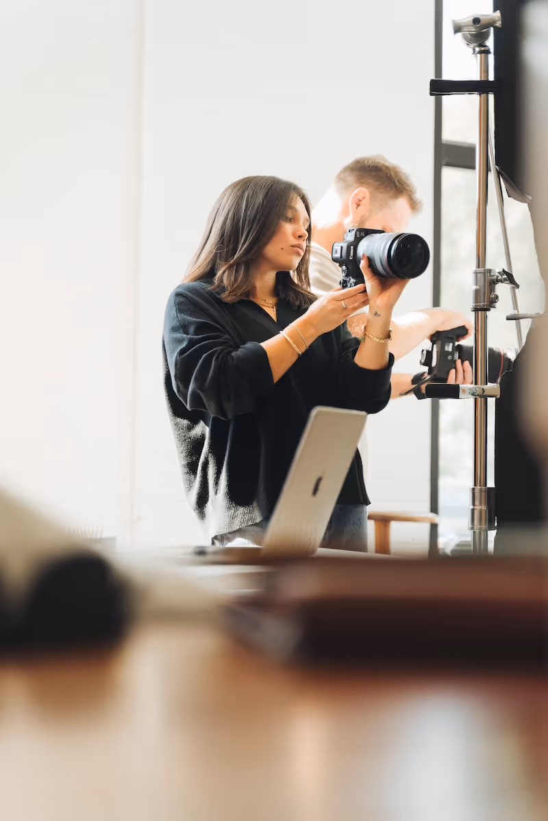 Woman in black sweater holding a DSLR camera adjusting camera settings in a bright room with a man working on equipment in the background.