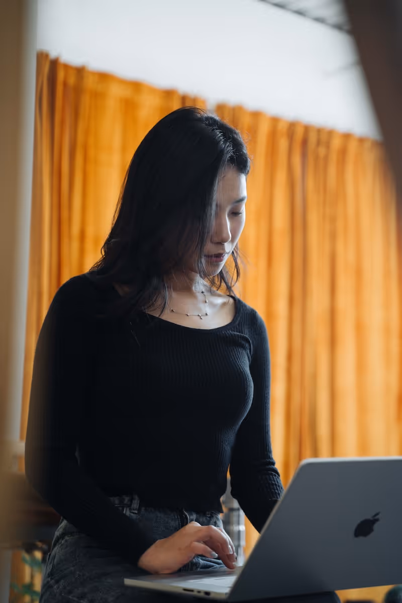 Woman with black hair and black top working on a silver Apple laptop in front of orange curtains.