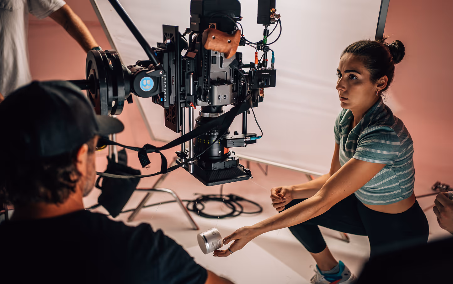 Woman in green striped shirt holding a jar in front of a professional film camera on a set with crew members.