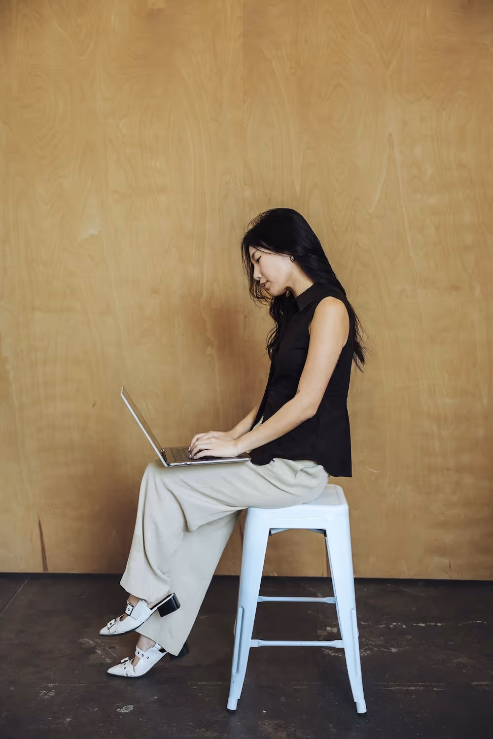Woman with long black hair wearing a black sleeveless top and beige pants sits on a white stool working on a laptop against a wooden wall.