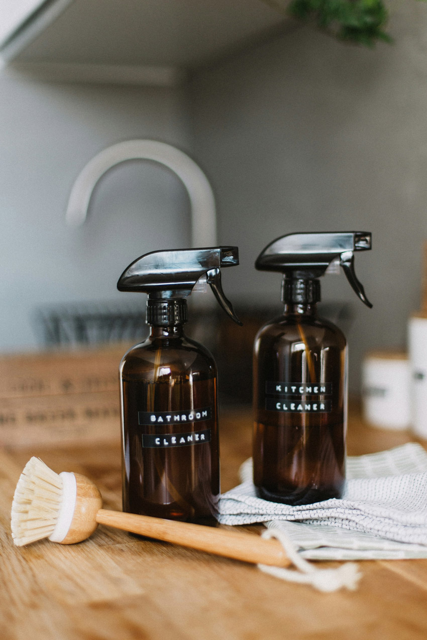Amber glass bottles labeled bathroom cleaner and kitchen cleaner on a countertop with a cleaning brush and cloth.