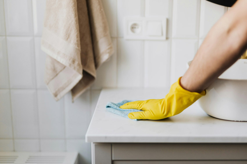 Person wearing yellow cleaning gloves wiping a bathroom counter with a microfiber cloth.