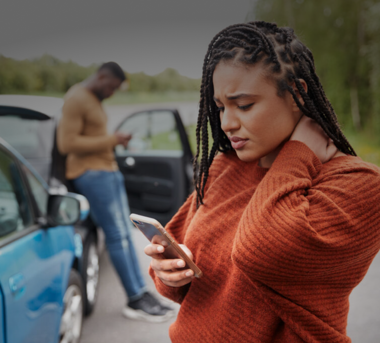 Woman holding her neck while looking at her phone near a car accident.