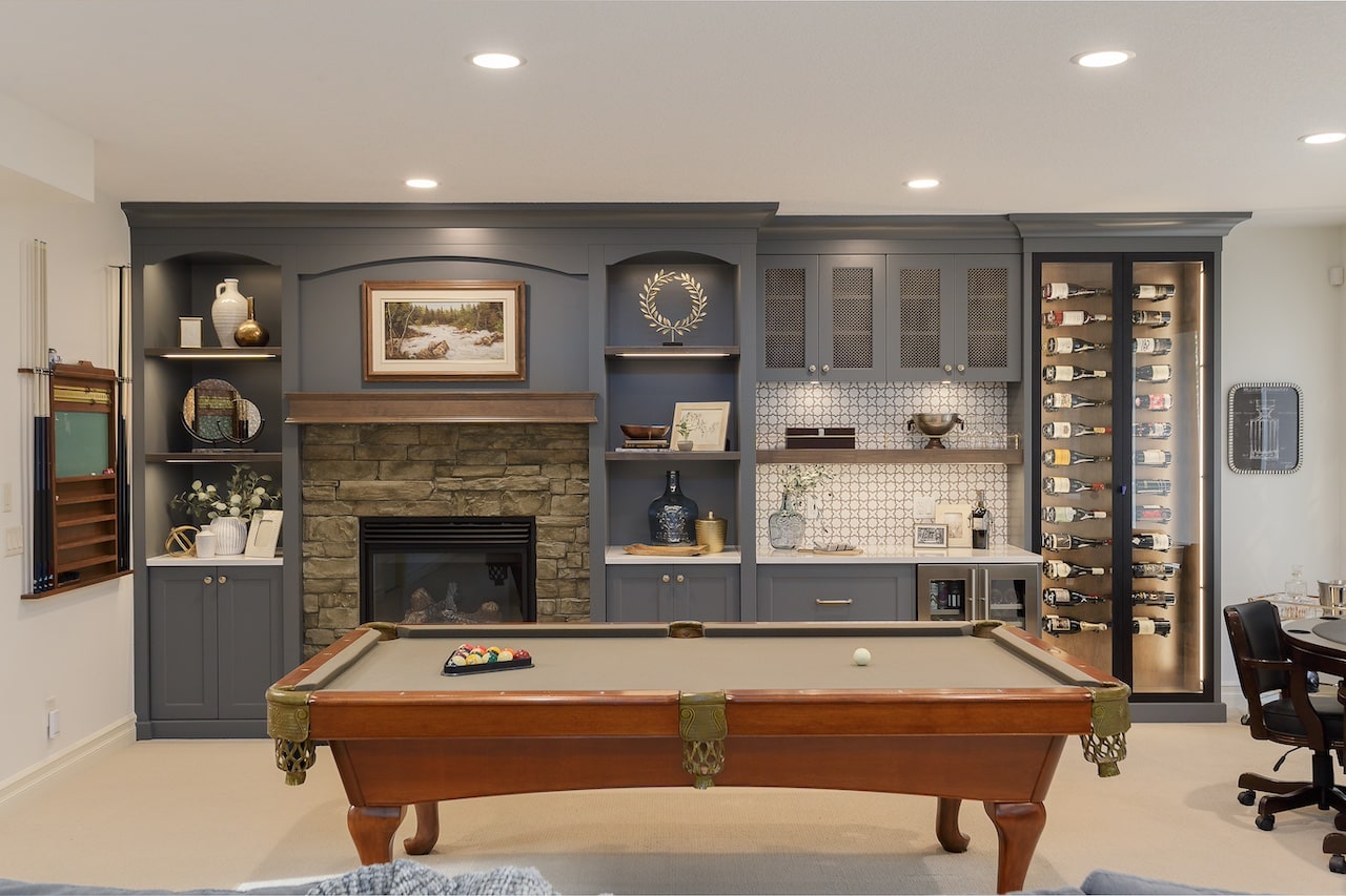 Game room with full-height custom built-ins, fireplace surround, and storage cabinetry in a warm grey finish. Cabinetry by Marvel Cabinetry.