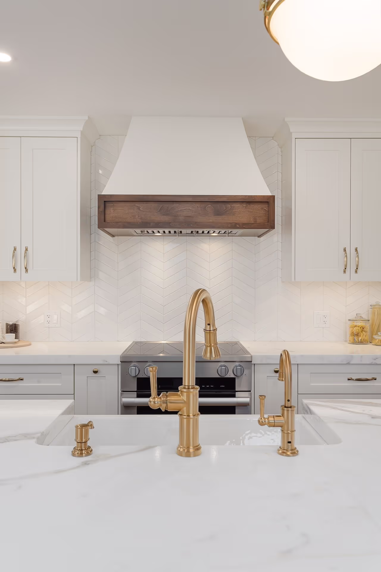 White kitchen with custom range hood, luxury cabinetry, gold faucet, and marble-style countertops. Cabinetry by Marvel Cabinetry.
