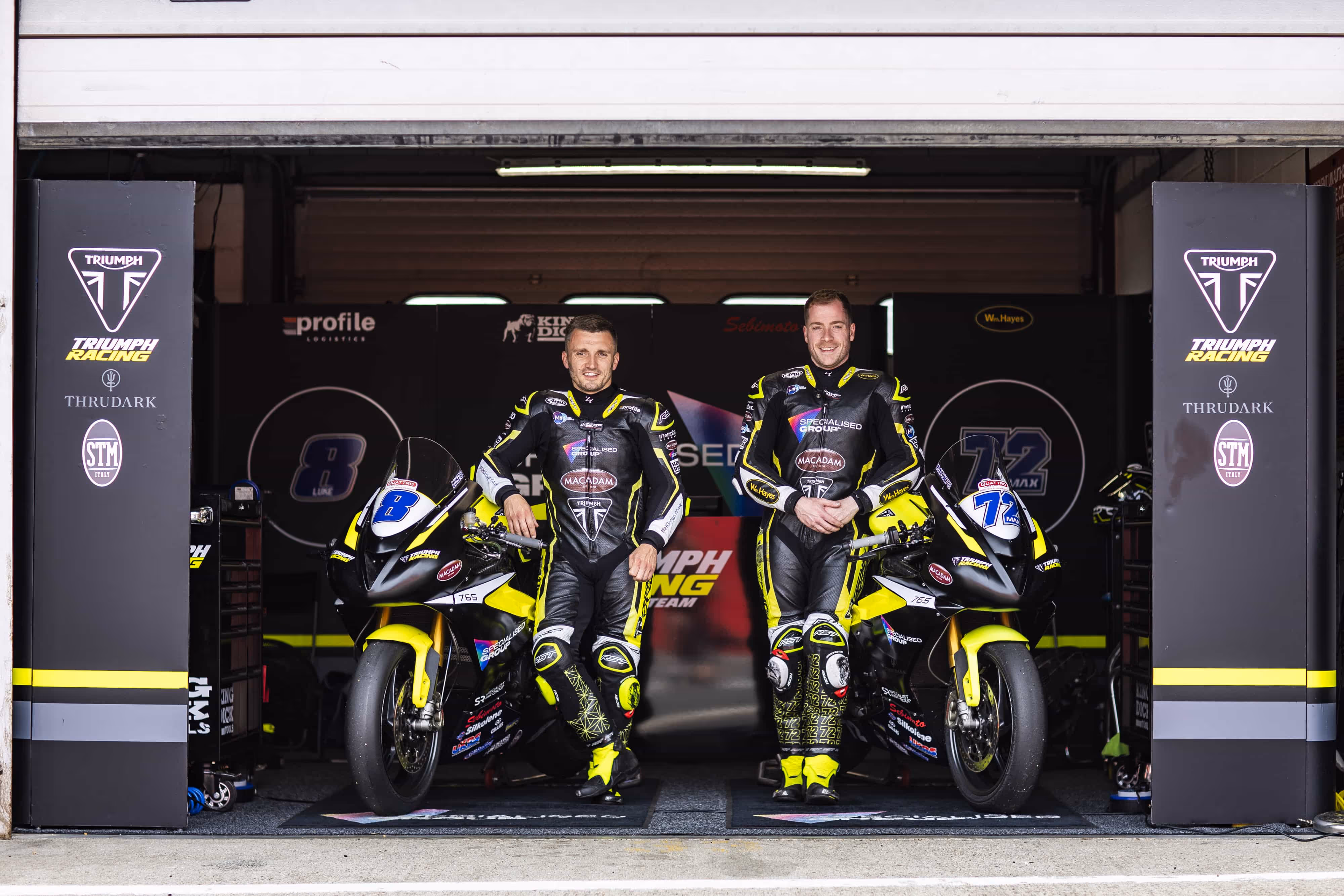 Two motorcycle racers in Triumph Racing gear posing with their bikes in a garage