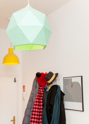 Ansicht eines Eingangberechs einer Wohnung mti einer gelben und einer grünen Lampe und einer Garderobe, in welcher Jacken und Schals hängen.