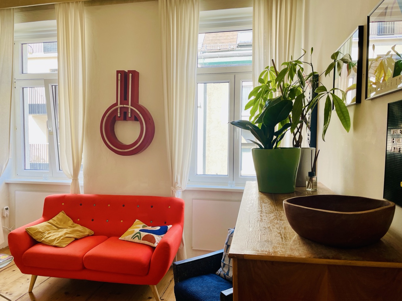 Ansicht eines eingerichteten Zimmers mit einer roten Couch und Holzkommode.
