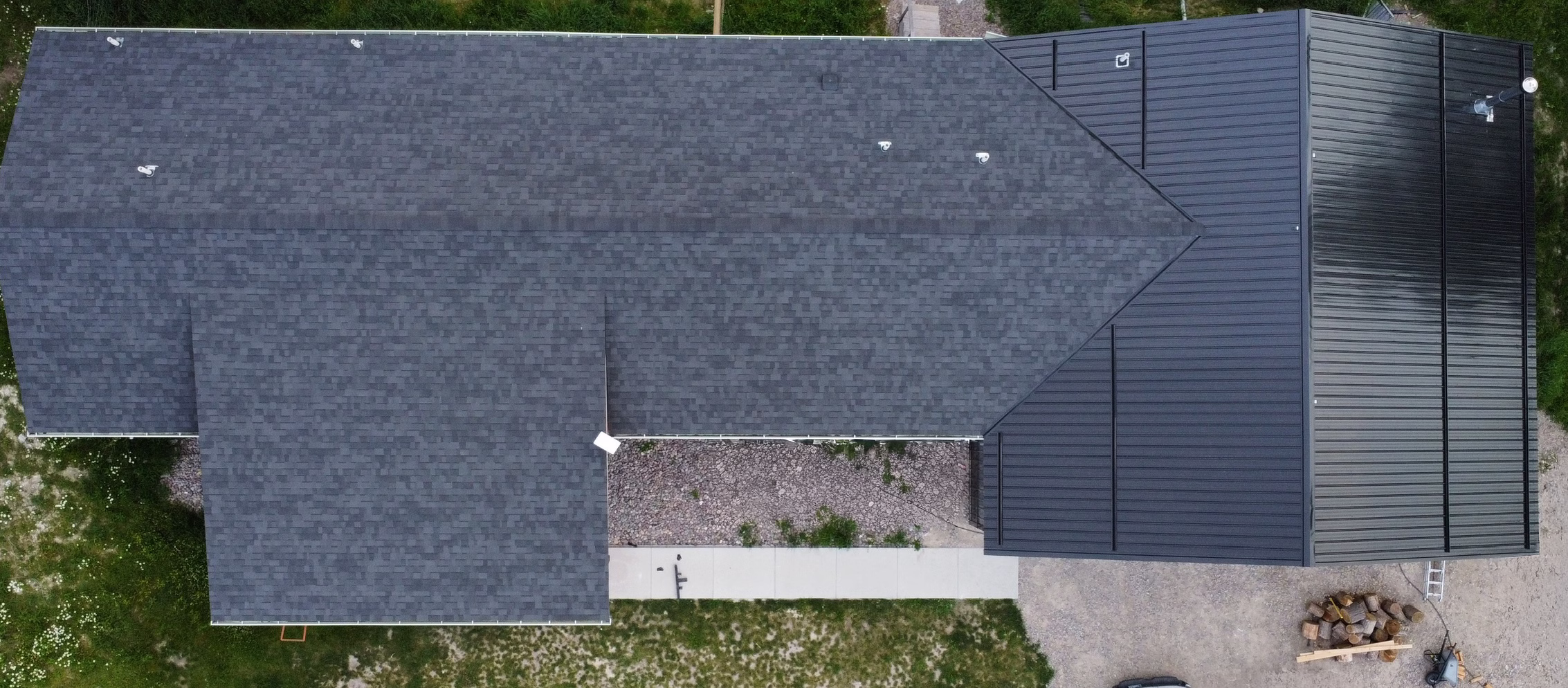 Overhead view of a house roof combining dark asphalt shingles and black metal roofing panels with surrounding grass and a small wood pile nearby.