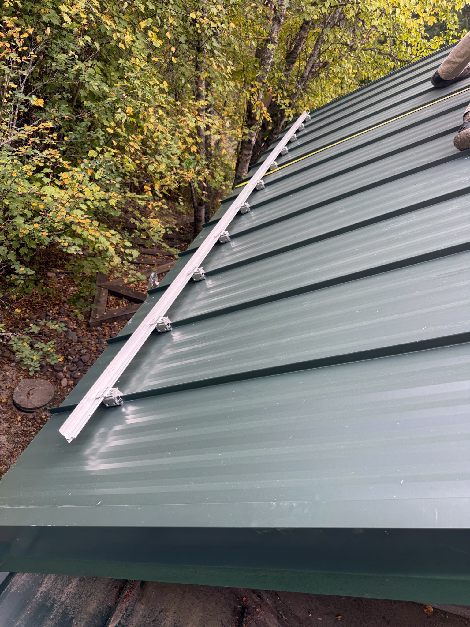 Green metal roof with mounted solar panel rails and a person’s boots visible at the top right near green trees.
