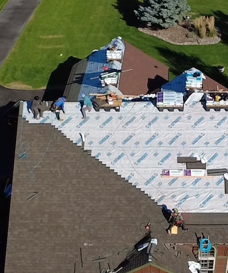 Aerial view of workers installing roof shingles on a house with roofing materials and tools spread out on the roof.