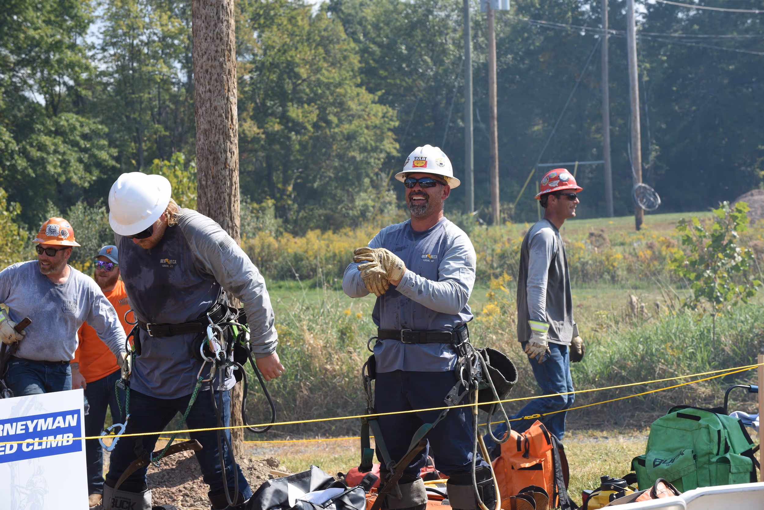 Lineworkers putting on safety gear and preparing equipment at an outdoor training or competition event, with trees and utility poles in the background.