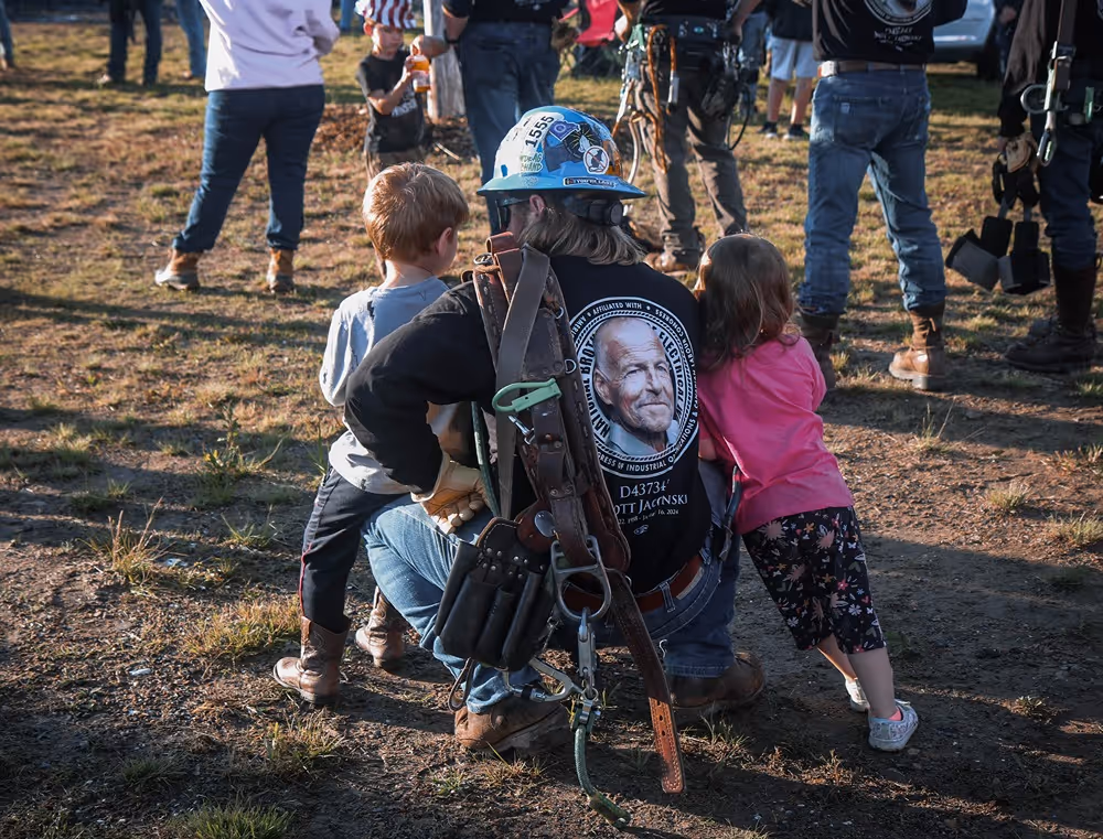 Lineworker kneeling with climbing gear as two young children hug him during a union event, highlighting family and community.