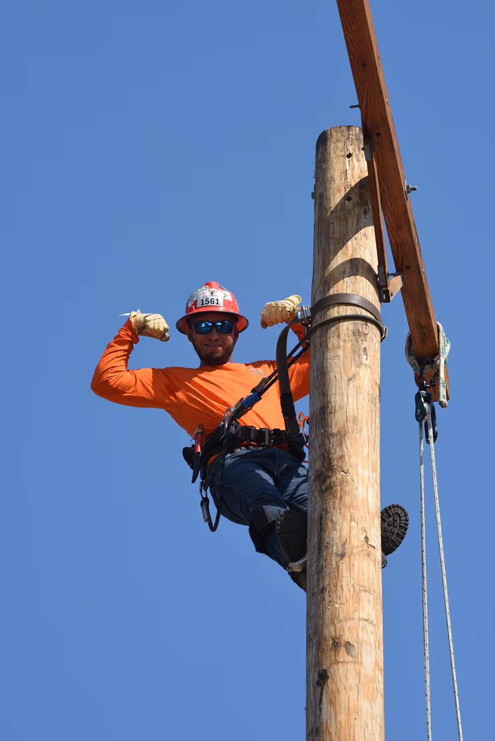 Lineworker wearing fall protection gear safely secured to a utility pole and flexing his arms against a clear blue sky.