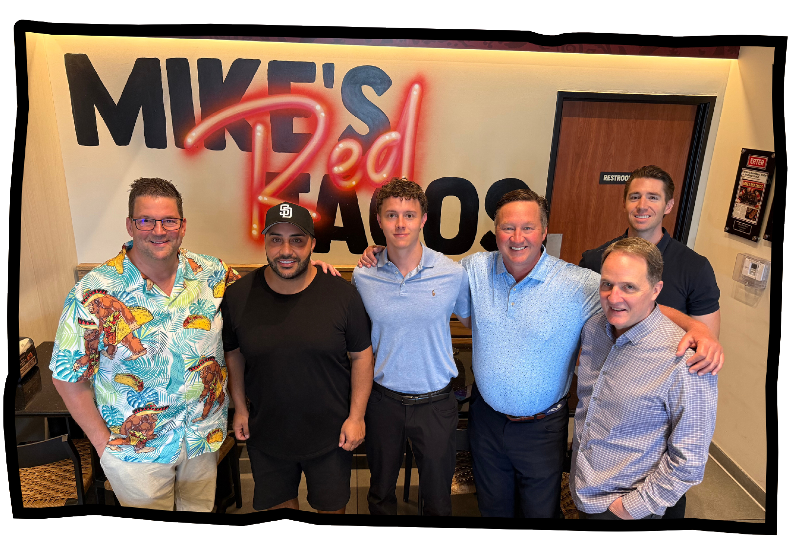 Six men standing and smiling inside Mike's Red Tacos restaurant, with the restaurant's logo on the wall behind them.