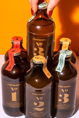 Hand holding Curious bottles numbered No.1, No.2, and No.3 on orange backdrop, beverage photography by Photos by Lars.