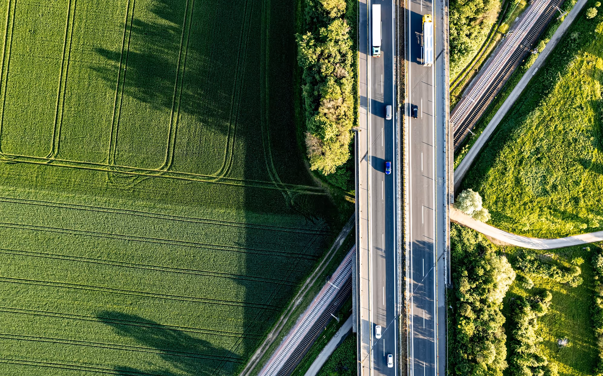 Aerial view of highway with several vehicles driving between green fields and railway tracks.