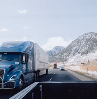 Blue semi-truck driving on a highway with snowy mountains in the background under a partly cloudy sky.