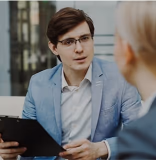 Man in light blue blazer and glasses holding a clipboard and talking with a woman in an office setting.