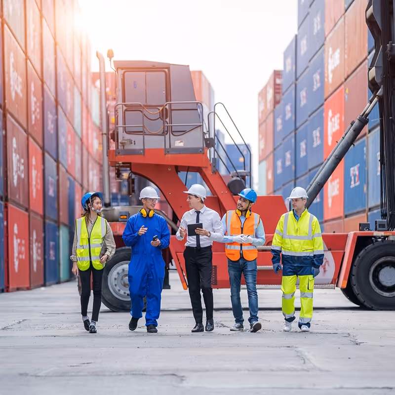Five workers wearing safety gear walking together at a shipping container yard with stacked containers and heavy machinery in the background.