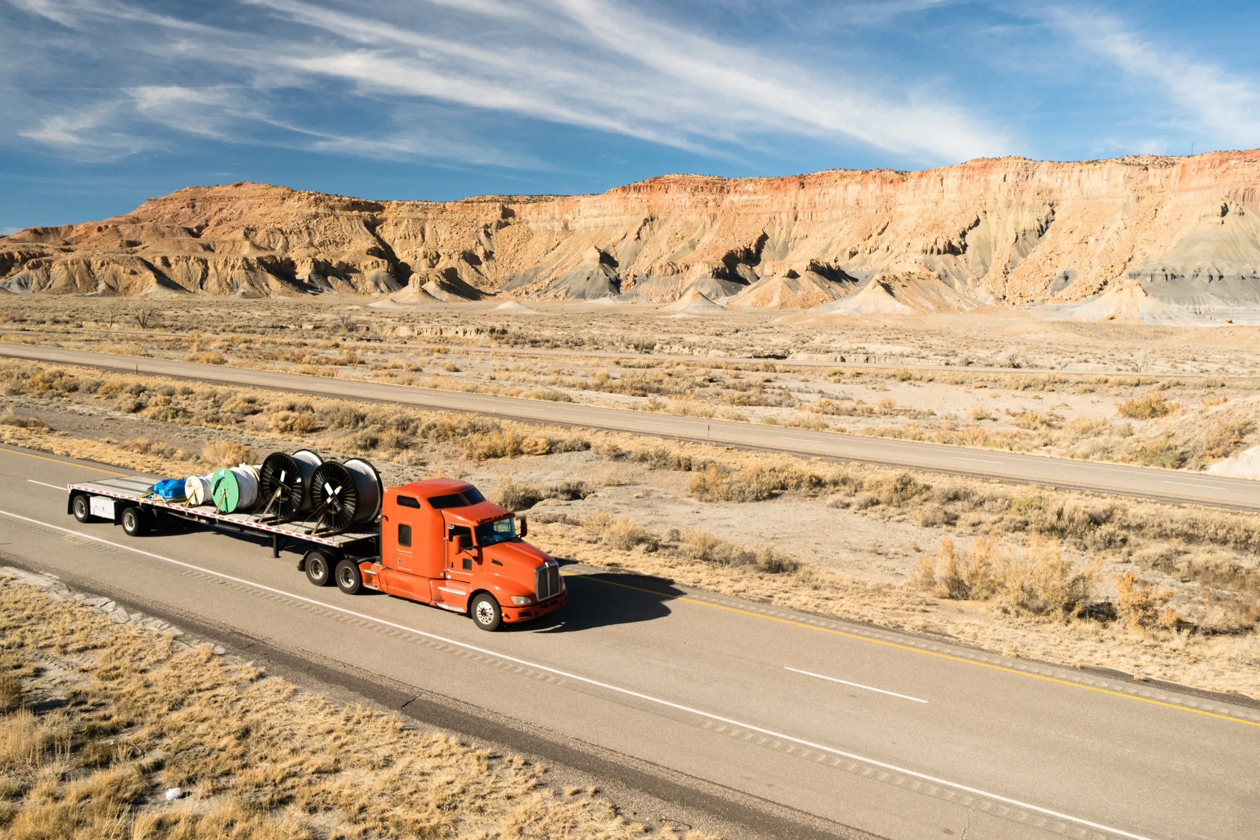 Red semi-truck hauling large cable reels on a flatbed trailer driving on a highway through a dry, rocky desert landscape.