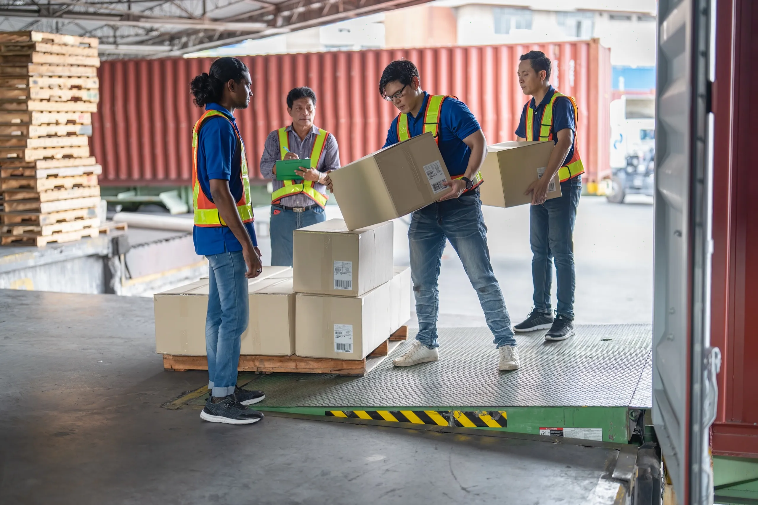 Four warehouse workers in safety vests loading cardboard boxes onto a pallet lift inside a shipping area.
